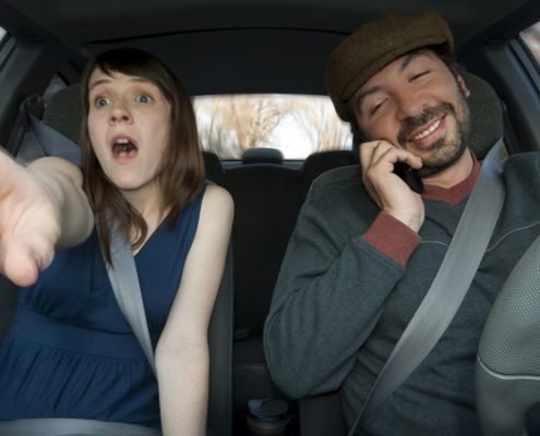 Ein Mann mit Handy am Steuer und eine Frau am Beifahrersitz, die erschrocken auf etwas zeigt