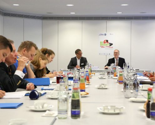 Konferenztisch mit Getränken, viele Personen am Tisch, 2 Wortführer am Tischende. Im Hintergrund Plakat der Kampagne Be Smart