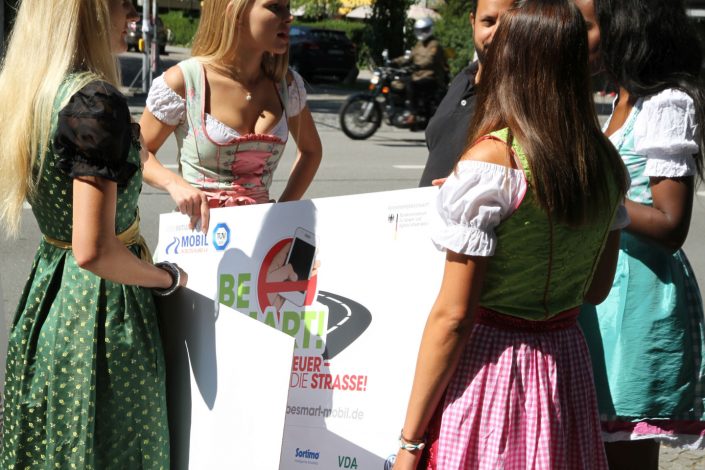 Vier Mädchen in Dirndl und Schildern in den Händen und ein Mann stehen vor einer belebten Straße im Kreis und reden
