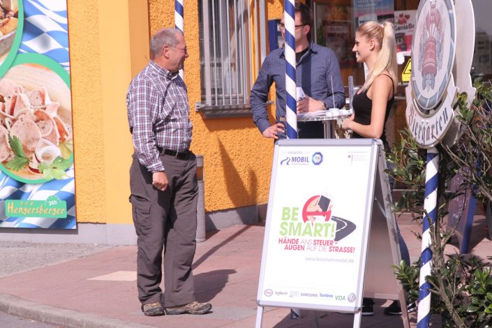 Ein Mann und eine Frau an einem Infostand sprechen mit einem interessierten älteren Herren. BeSmart Plakat im Vordergrund