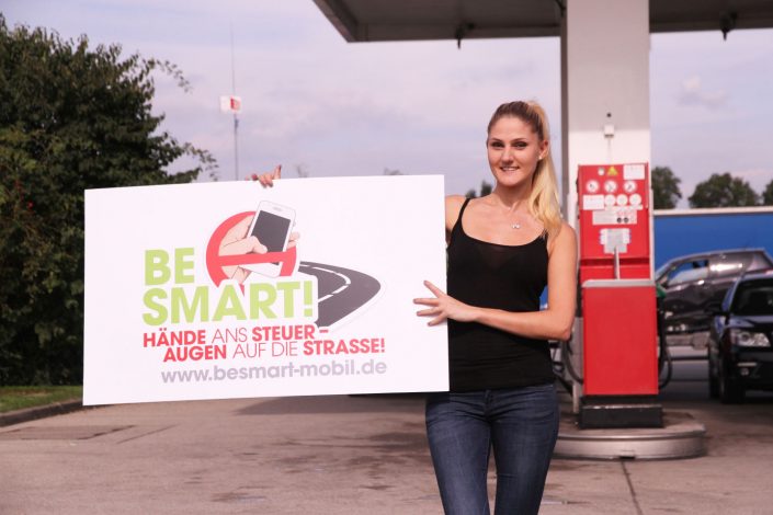 Blonde junge Frau mit BeSmart Plakat vor Esso-Tankstelle