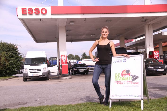 Blonde junge Frau mit BeSmart Plakat vor Esso-Tankstelle