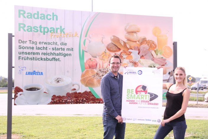 Mann und Frau mit BeSmart Plakat vor einem Rasthofschild