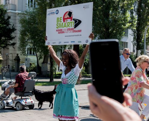 Ein junges Mädchen in Dirndl hält auf einer belebten Straße ein Schild in die Höhe, ein Schwarzes Smartphone im Vordergrund