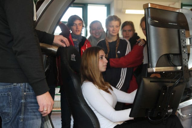 Mehrere Jugendliche stehen um Fahrsimulator herum während ein Mädchen fährt