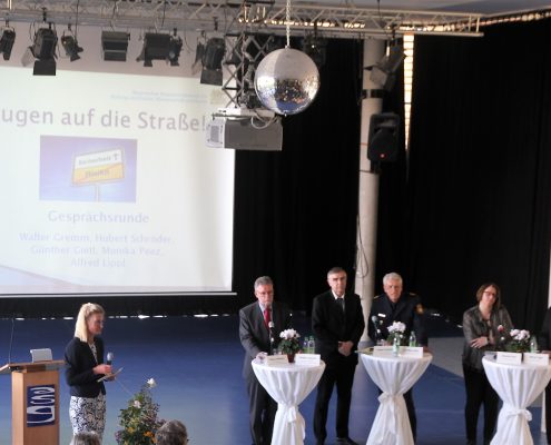 Fünf Erwachsene stehen an Stehtischen in einer Aula und eine Frau hält ein Mikrofon in der Hand