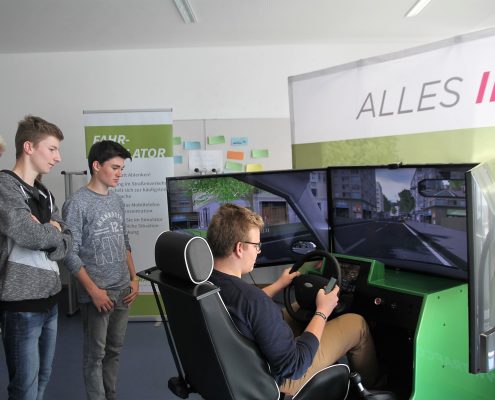 Ein Junge der mit dem Fahrsimulator fährt und drei weitere Jungs die zusehen und links von ihm stehen